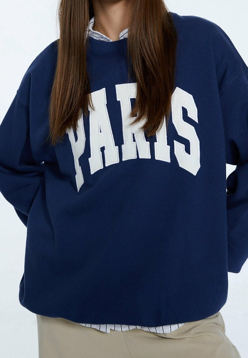 Granatowy sweatshirt wykonany z miękkiej tkaniny, z dużą białą grafiką "PARIS" na przodzie oraz luźnym krojem.