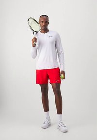 Biela košeľa s dlhým rukávom a gombíkovým golierom, červené športové šortky, drží tenisovú raketu a loptičku, na nohách biele tenisky s čiernymi detailmi.