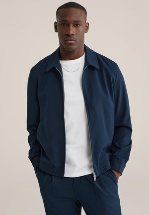 Egy férfi sötétkék cipzáras dzsekit visel fehér póló fölött, sötétkék nadrágot, zsebében tartja a kezét, bézs háttér előtt áll.