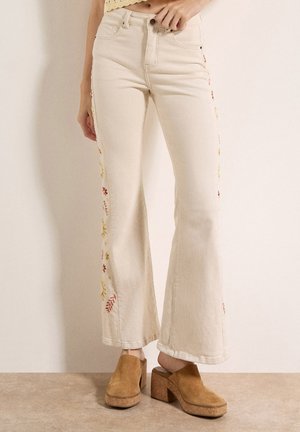 Donna che indossa jeans a vita alta color crema svasati con ricami floreali ai lati e zoccoli platform in camoscio color tan su pavimento beige.