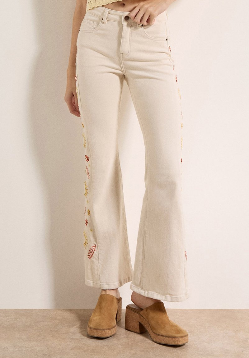 Donna che indossa jeans a vita alta color crema svasati con ricami floreali ai lati e zoccoli platform in camoscio color tan su pavimento beige.