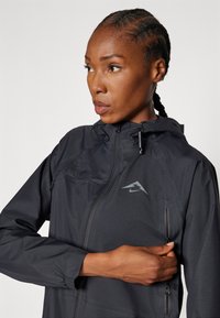 Veste légère noire avec capuche, dotée d'une poche poitrine zippée, d'un tissu texturé et d'un logo réfléchissant sur le côté gauche.