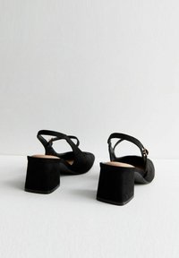 Zapatos de tacón de ante negro tipo slingback con tacones gruesos y correas de tobillo ajustables, mostrados desde la parte posterior sobre una superficie blanca.