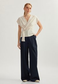 Top avvolgente color crema con nodo in vita, abbinato a pantaloni larghi a righe navy. Tessuto liscio, design moderno.