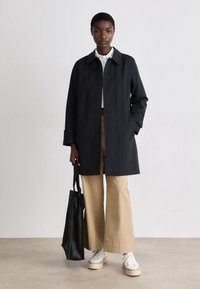 Abrigo negro de tipo trench con cuello, usado sobre una camisa blanca y pantalones beige. El modelo lleva un bolso de mano negro y calza zapatillas blancas.