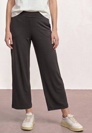 Personne portant un pantalon noir ample, des baskets blanches avec des semelles beiges et un haut blanc rentré, debout devant un fond clair.
