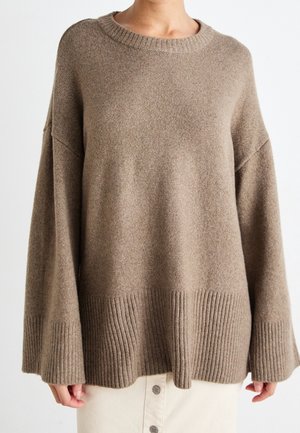 Femme portant un pull en maille marron ample avec des poignets et un ourlet larges côtelés, associé à une jupe boutonnée de couleur claire.