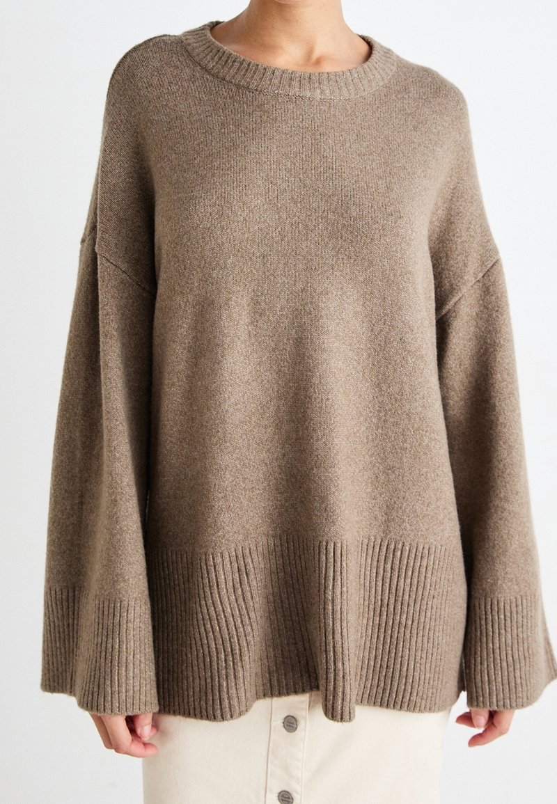 Femme portant un pull en maille marron ample avec des poignets et un ourlet larges côtelés, associé à une jupe boutonnée de couleur claire.