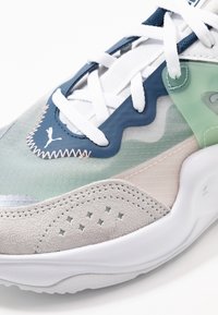 Sneakers Puma realizzati con una combinazione di materiali blu, verdi e grigi, sovrapposizioni testurizzate e lacci bianchi. Elementi di design perforati degni di nota.