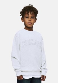 Sweatshirt gris avec "CHICAGO" brodé dans une couleur contrastante. Présente un col rond et une coupe décontractée. Porté sur une chemise blanche.