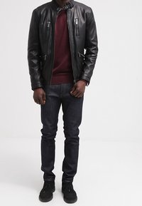 Schwarze Lederjacke mit hohem Kragen, mehreren Reißverschlusstaschen, kombiniert mit einem kastanienbraunen Pullover und dunklen Denim-Jeans. Schwarze Schuhe runden das Outfit ab.