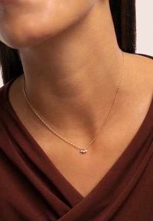 Goldene Kette mit zwei sternförmigen Anhängern, die mit kleinen transparenten Steinen besetzt sind, getragen über einem braunen Stoffoberteil.