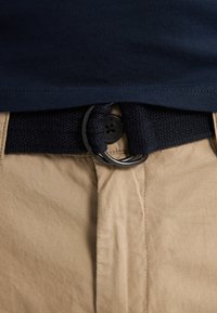 Curea împletită de culoare bleumarin cu o cataramă metalică rotundă, asortată cu pantaloni khaki. Țesătura este netedă, cu un ușor luciu și o croială obișnuită.