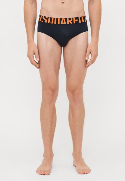 Modello maschile che indossa slip neri con ampia vita arancione, su cui è presente un grande testo parzialmente visibile, su uno sfondo bianco semplice.