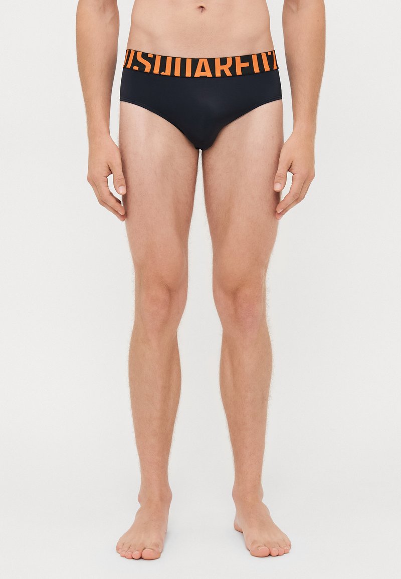Männliches Modell, das schwarze Slip mit breitem orangefarbenem Bund trägt, auf dem eine große teilweise sichtbare Schrift auf einem schlichten weißen Hintergrund zu sehen ist.