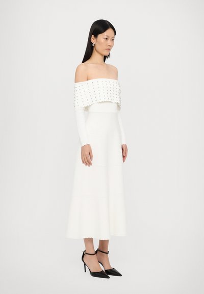 Witte off-shoulder jurk met een gestructureerd bovenstuk, een golvende zoom en lange mouwen. Zwarte hoge hakken met bandjes complementeren de outfit.