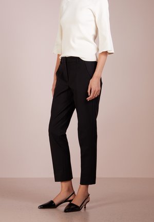 Kobieta w czarnych odsłoniętych spodniach, białym topie z rękawami do łokcia oraz czarnych butach na niskim obcasie z paskiem za piętą stoi na beżowej podłodze.