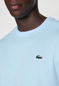 T-shirt di cotone azzurro chiaro con scollo rotondo e maniche corte, dotata di un piccolo logo di coccodrillo verde e rosso sul petto.