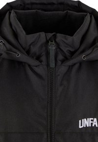 Svart jakke med glidelås og justerbar hette, med glatt stoff, fleecefôret innside, og hvit "UNFA"-logo på forsiden.