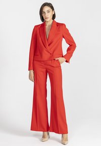 Czerwony, dopasowany garnitur z gładkiej tkaniny; składa się z marynarki dwurzędowej z klapami szpiczastymi oraz spodni o szerokich nogawkach. Stylizację dopełniają neutralne szpilki.