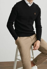 Pull noir en maille à col en V avec une surface texturée et un petit logo ; assorti à un pantalon chino beige et des baskets blanches, assis sur un tabouret blanc.
