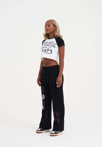 Czarny i biały przycięty T-shirt z grafiką, zestawiony z luźnymi czarnymi spodniami z fioletowymi akcentami i wzorami. Stylizowany z trampkami.