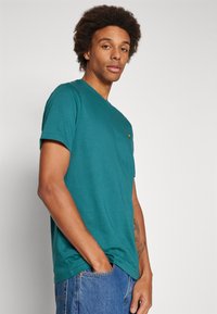 T-shirt teal a maniche corte con collo rotondo, dotato di un piccolo logo ricamato sul lato sinistro superiore, realizzato in un tessuto morbido e liscio.