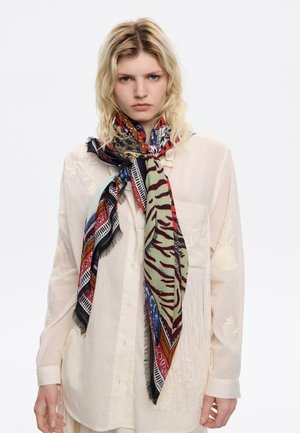 Donna bionda che indossa una blusa ricamata color crema e trasparente e una grande sciarpa multicolore annodata attorno al collo.