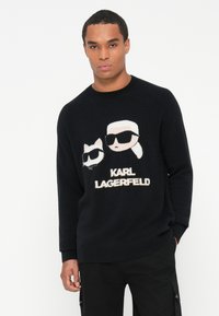 Fekete kötött pulóver, amelyen két napszemüveget viselő karakter grafikája látható, alatta a "KARL LAGERFELD" szöveg krémszínben. Hosszú ujjú, kerek nyakú.