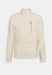 Pullover in pile color crema con colletto a mezza zip. Presenta una tasca frontale e una tasca sul petto con zip a contrasto e dettagli rossi.