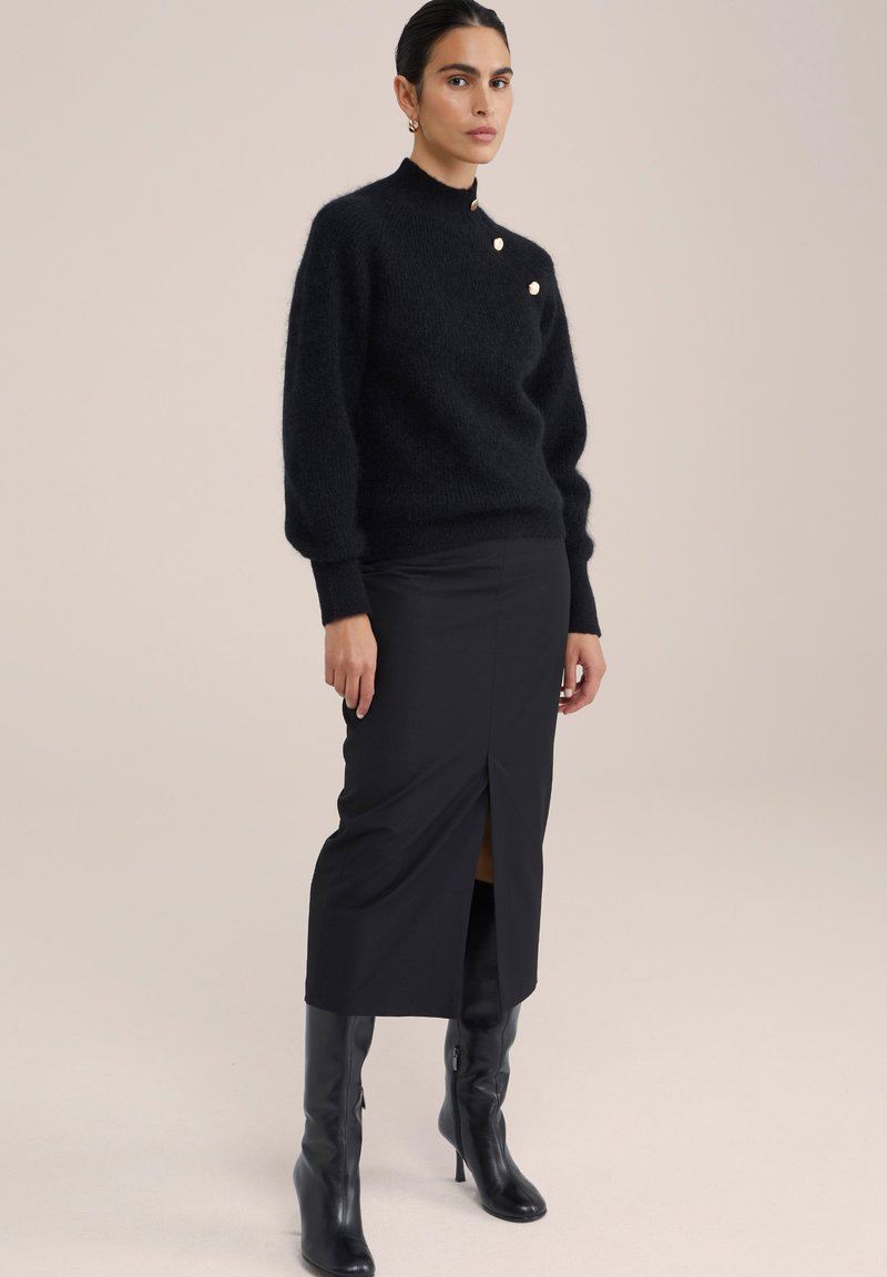 Vrouw die een zwarte fuzzy trui met drie gouden knopen, een zwarte midi-rok met een split aan de voorkant en zwarte hoge laarzen met hakken tot de knie draagt.