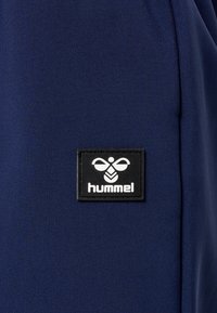 Marineblauem Textil mit glatter Textur, das ein rechteckiges schwarzes Patch zeigt, auf dem das "hummel"-Logo in Weiß abgebildet ist.