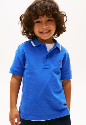 Jeune enfant aux cheveux bouclés souriant, portant un polo à manches courtes bleu avec des bordures blanches et un pantalon foncé, debout devant un fond blanc.