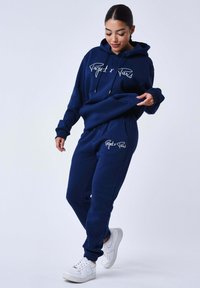 Conjunto de sudadera con capucha y pantalones deportivos azul marino hecho de tela suave, con un logo blanco en ambas prendas, y puños y cintura acanalados.