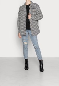 Schwarz-weiße Hahnentwist-Hemdenjacke mit zwei Fronttaschen, getragen über einem schwarzen Pullover, kombiniert mit zerrissenen hellblauen Jeans und schwarzen Stiefeletten.