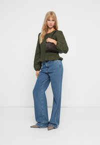 Blouse smock olive verte avec manches longues, associée à un jean bleu à jambes larges et un petit sac à main sombre. Des talons aiguilles beiges complètent le look.