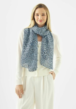 Vrouw die een witte cardigan en broek draagt met een blauwe en zwarte luipaardprint sjaal, glimlachend, hand in de zak, op een witte achtergrond.