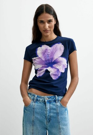 Donna che indossa una t-shirt blu navy con una grande stampa floreale viola e bianca, abbinata a jeans in denim azzurro chiaro a vita alta.