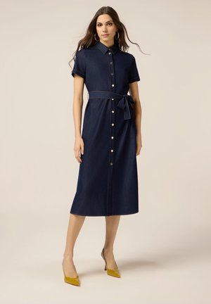 Denim midi-jurk met korte mouwen, knopen aan de voorkant en een strikceintuur. Donkerblauw met contrasterende stiksels, gecombineerd met gele puntige hakken.
