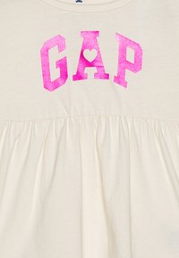 Kremfarget T-skjorte med rynket bunn, med stor rosa glitrende "GAP"-tekst og et hjerte-design i sentrum.
