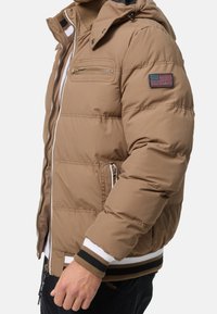 Beige Pufferjacke mit Reißverschlusstaschen und einem Aufnäher der amerikanischen Flagge auf dem linken Ärmel, getragen über einem weiß-schwarz gestreiften, umgeschlagenen Pullover.