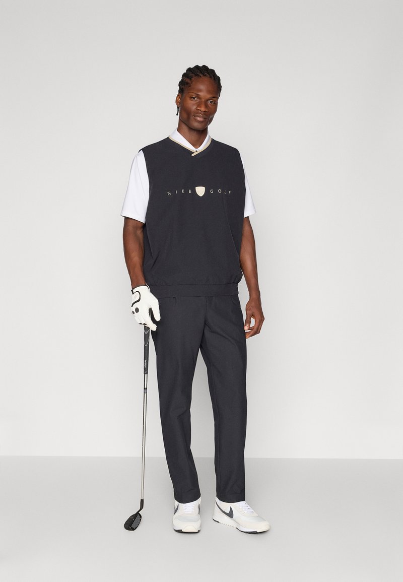 Tenue de golf Nike comprenant un pull sans manches noir avec logo, une chemise blanche à manches courtes, un pantalon gris et des baskets blanches ; tenant un club de golf.