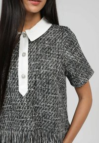 Chemise noire et blanche à manches courtes avec un motif texturé, col blanc, et deux boutons décoratifs. Détail de franges à l'ourlet.