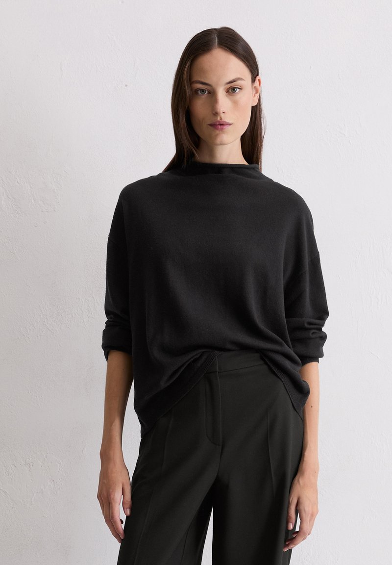 Schwarzer, übergroßer Pullover mit hohem Kragen und umgeschlagenen Ärmeln. Kombiniert mit einer taillierten schwarzen Hose, beide haben eine weiche Textur und ein einfaches Design.