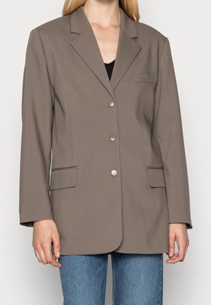 Frau trägt taupefarbenen, einreihigen Blazer mit Kerbkragen, drei Knöpfen, Klappentaschen und blauen Jeans vor schlichtem Hintergrund.