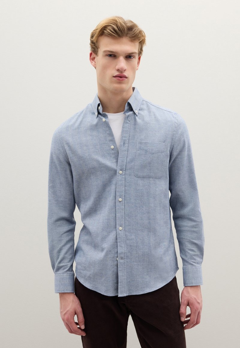 Camicia azzurro chiaro con bottoni, realizzata in tessuto morbido, con taschino sul petto, maniche lunghe e orlo arrotondato. Abbinata a pantaloni marrone scuro.