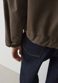 Bruine, lichtgewicht jas met een gladde textuur en aansluitende mouwen, gecombineerd met donkerblauwe denimjeans met oranje stikseldetails.