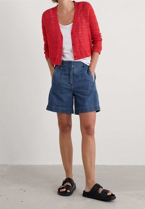 Personne portant un cardigan en maille rouge par-dessus un top blanc, un short en jean bleu taille haute et des sandales noires à enfiler, debout sur un sol gris.