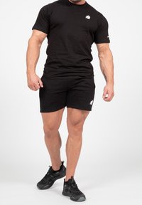 Černá sportovní souprava obsahující tričko s krátkým rukávem a šortky. Texturovaná tkanina, přiléhavý design a bílé akcenty s logem. Sportovní boty v černé barvě.