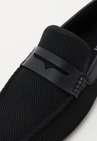 Chaussure noire à enfiler avec une tige en maille, une sangle en cuir, une surface texturée et un design minimaliste. Présente un détail de ligne lisse sur la sangle.